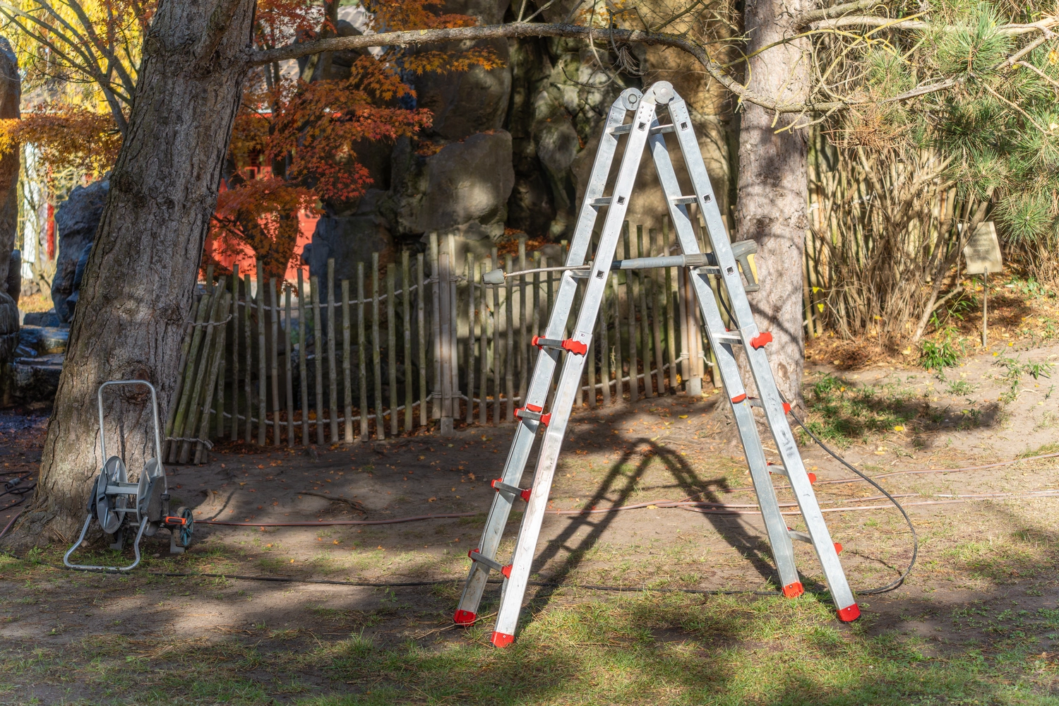 R.S Sicco UG – Wasser- & Brandschadensanierung, Reinigung und Gebäudedienste - Leiter unter Bäumen. Eine A-förmige Leiter steht für Baumarbeiten neben einer Schlauchtrommel. Sonnenlicht durchflutet das Herbstlaub umgibt die Baustelle.
